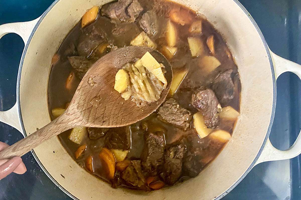 A wooden spoon holding softened vegetables cooked in Spicy Beef Stew.