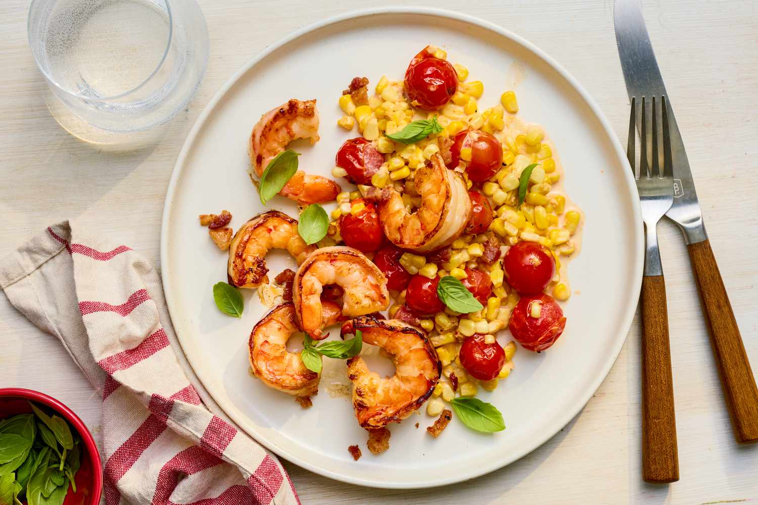Plate of creamy skillet shrimp with corn, pancetta, and tomatoes at a table setting with a glass of water, a white and red table napkin on the counter, and a bowl of basil