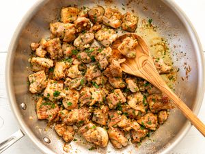 Chunks of cooked chicken in a skillet with herbs and spices, accompanied by a wooden spoon