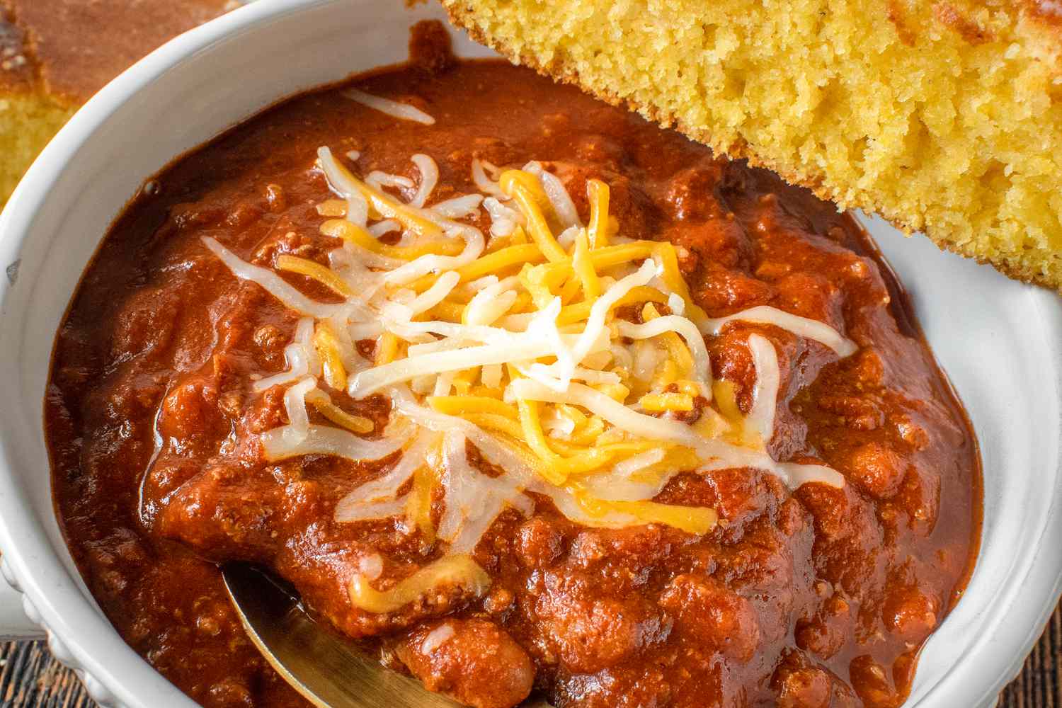 Closeup view of a white bowl of chili with cheese topping and spoon along with a slice of cornbread balanced on the edge of the bowl