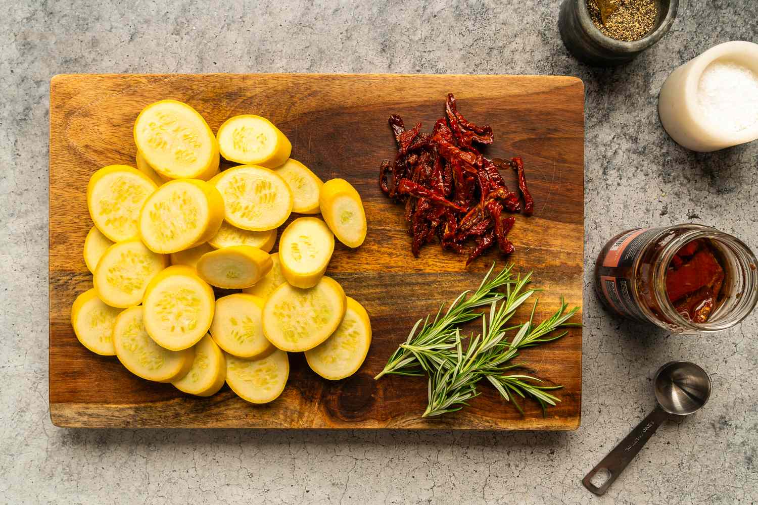 Slices of zucchini on a wooden cutting board accompanied by rosemary sprigs and sundried tomatoes with seasonings nearby