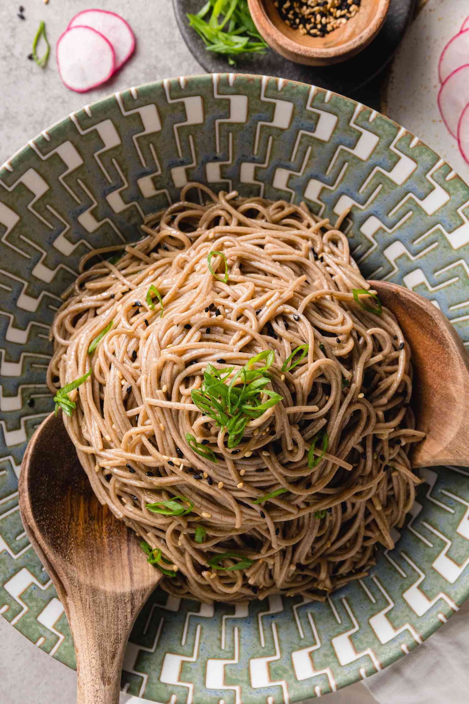 Sesame Soba Noodles Garnished with Spring Onions in a Bowl with Wooden Spoons Next to Plate with Spring Onions and Seasoning 