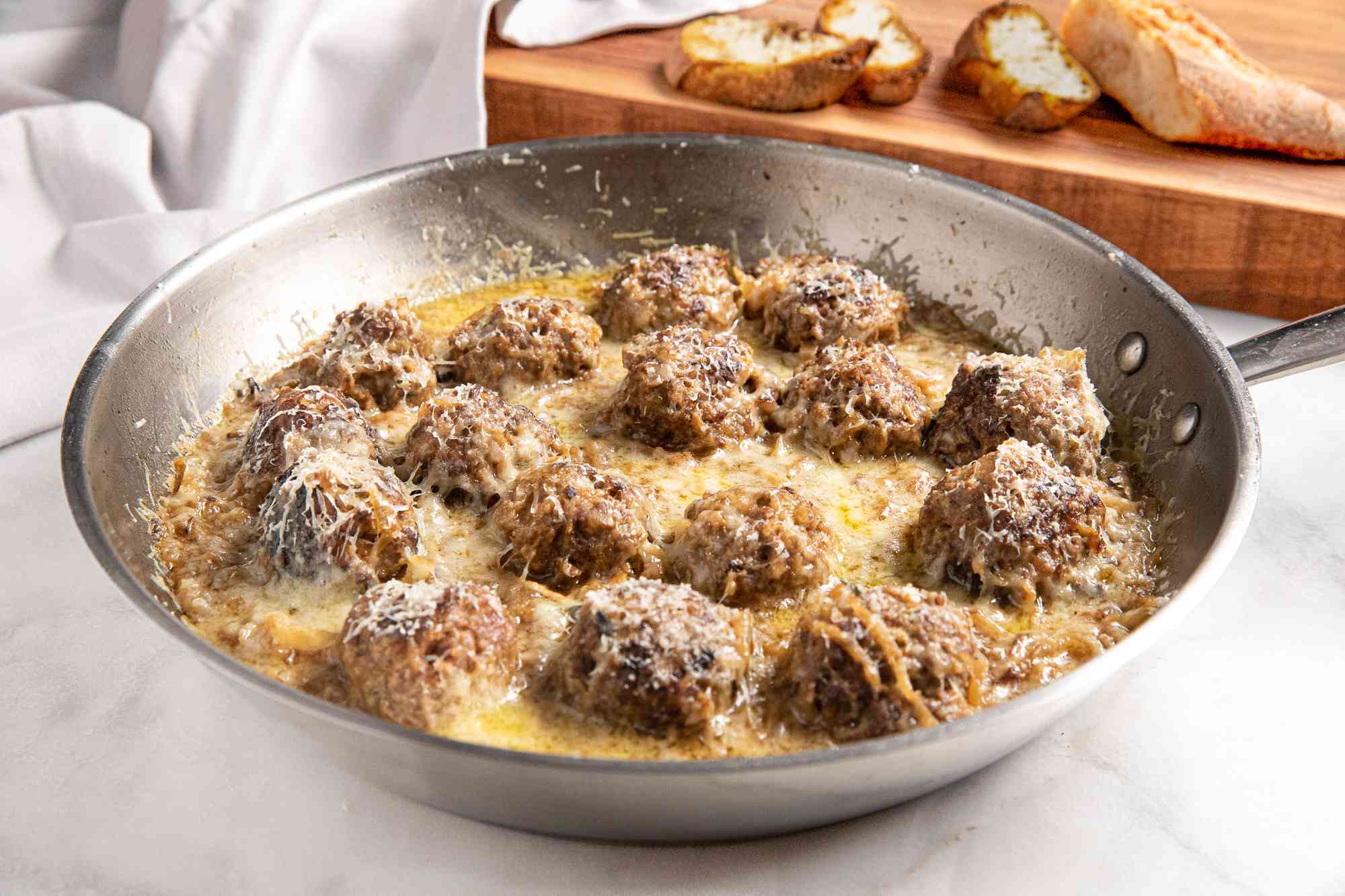 French Onion Meatballs in a Pan, and in the Background, Toasted Baguette Slices on a Wooden Platter and a White Table Cloth Draped Onto It