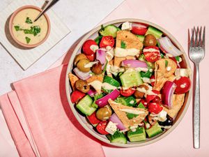 Bowl of salad topped with hummus salad dressing, and in the surroundings, a bowl of hummus salad dressing on a white coaster and a pink table napkin