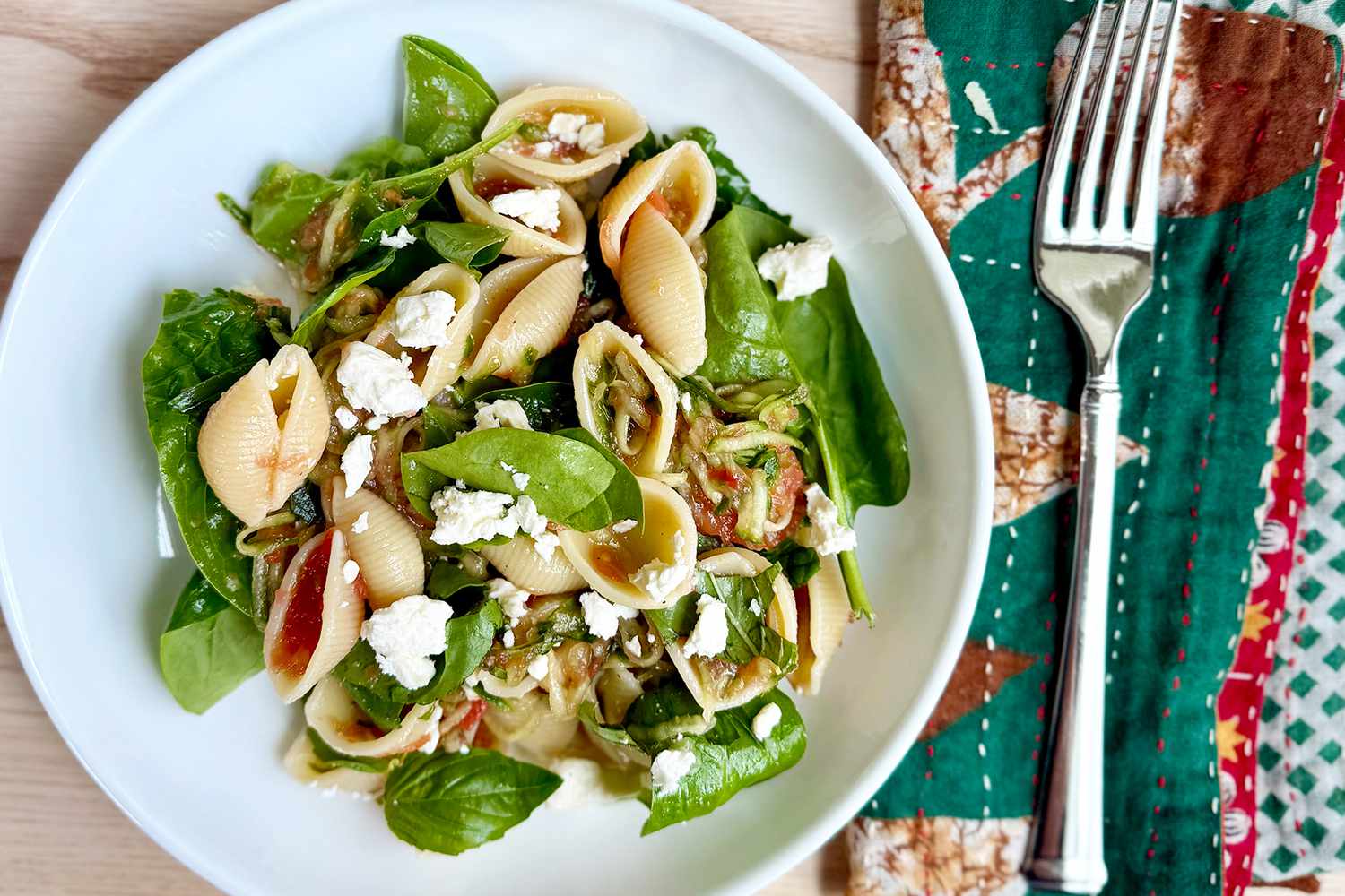 A plate of pasta shells with fresh vegetables and herbs, garnished with crumbled cheese, next to a fork on a colorful napkin