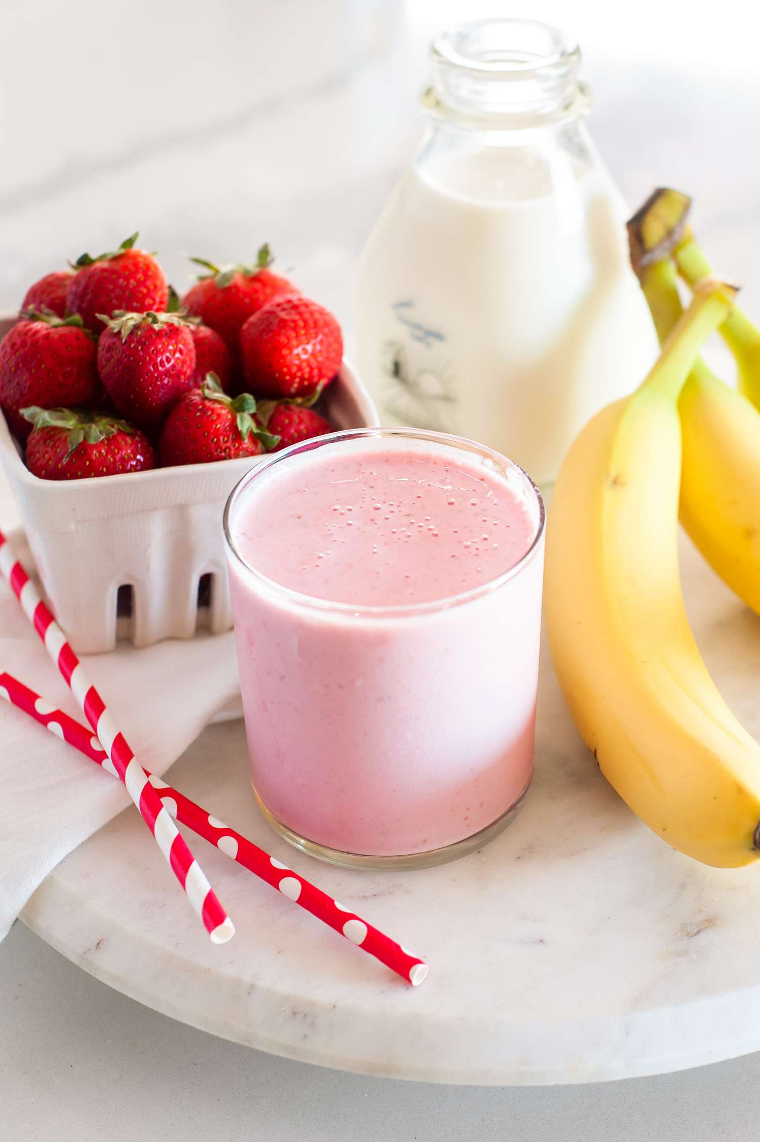 Strawberry banana smoothie in a glass with ingredients of milk, bananas, and strawberries nearby