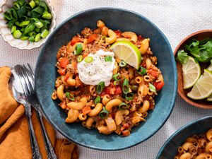 Two Bowls of One-Pot Taco Pasta Topped With Sour Cream, Sliced Scallions and a Lime Wedge, and in the Surroundings, a Bowl With Lime Wedges, a Bowl With Sliced Green Onions, a Bowl With Sour Cream, Glasses With a Drink, and a Mustard Colored Table Napkin With a Pair of Silverware