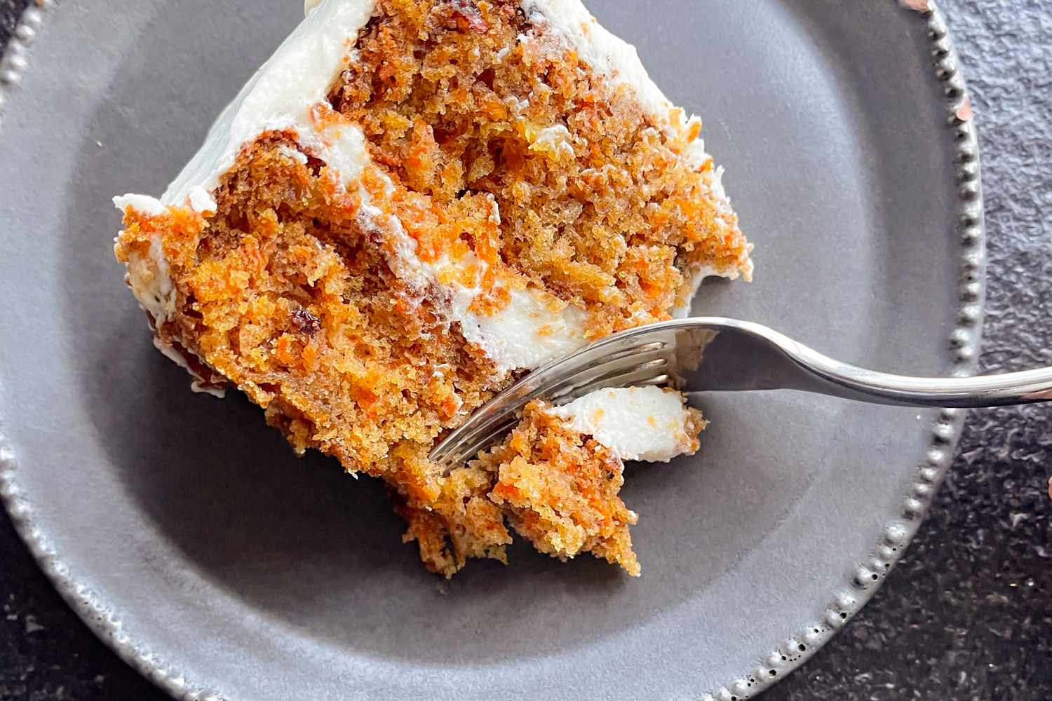 A carrot cake slice on a plate with a fork cutting into the cake
