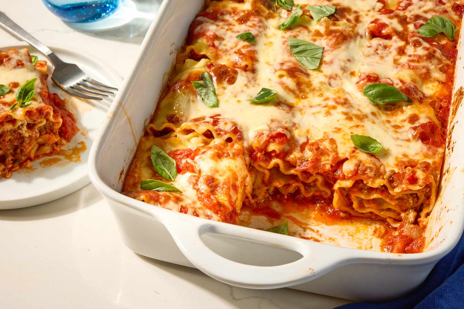 Baked lasagna rollups in a dish topped with melted cheese and basil leaves with a serving on a plate nearby