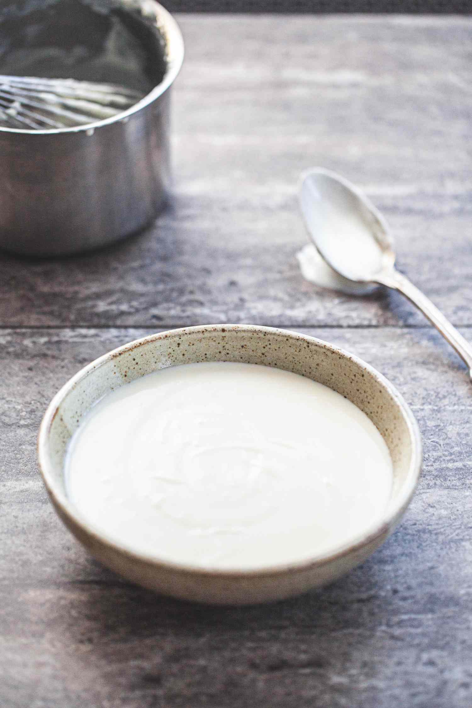 Side view of basic béchamel sauce in a bowl with a spoon and sauce pan above it.