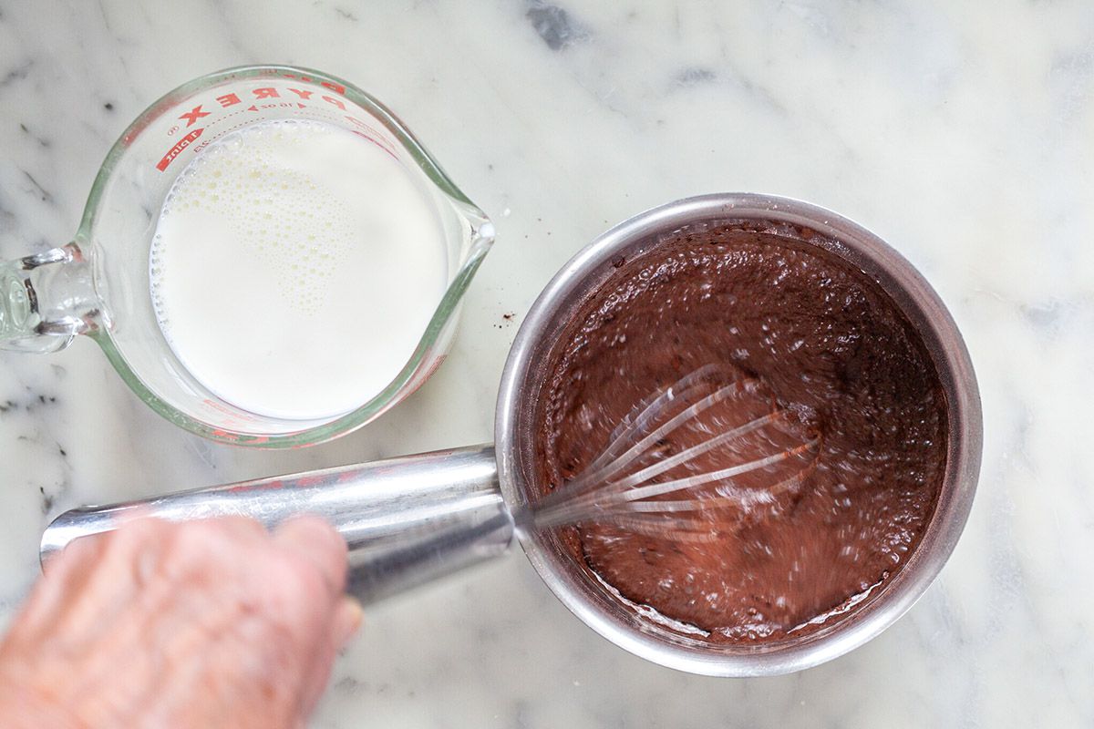 Whisking melted chocolate in a pan and a glass measuring cup of cream to make chocolate pudding.