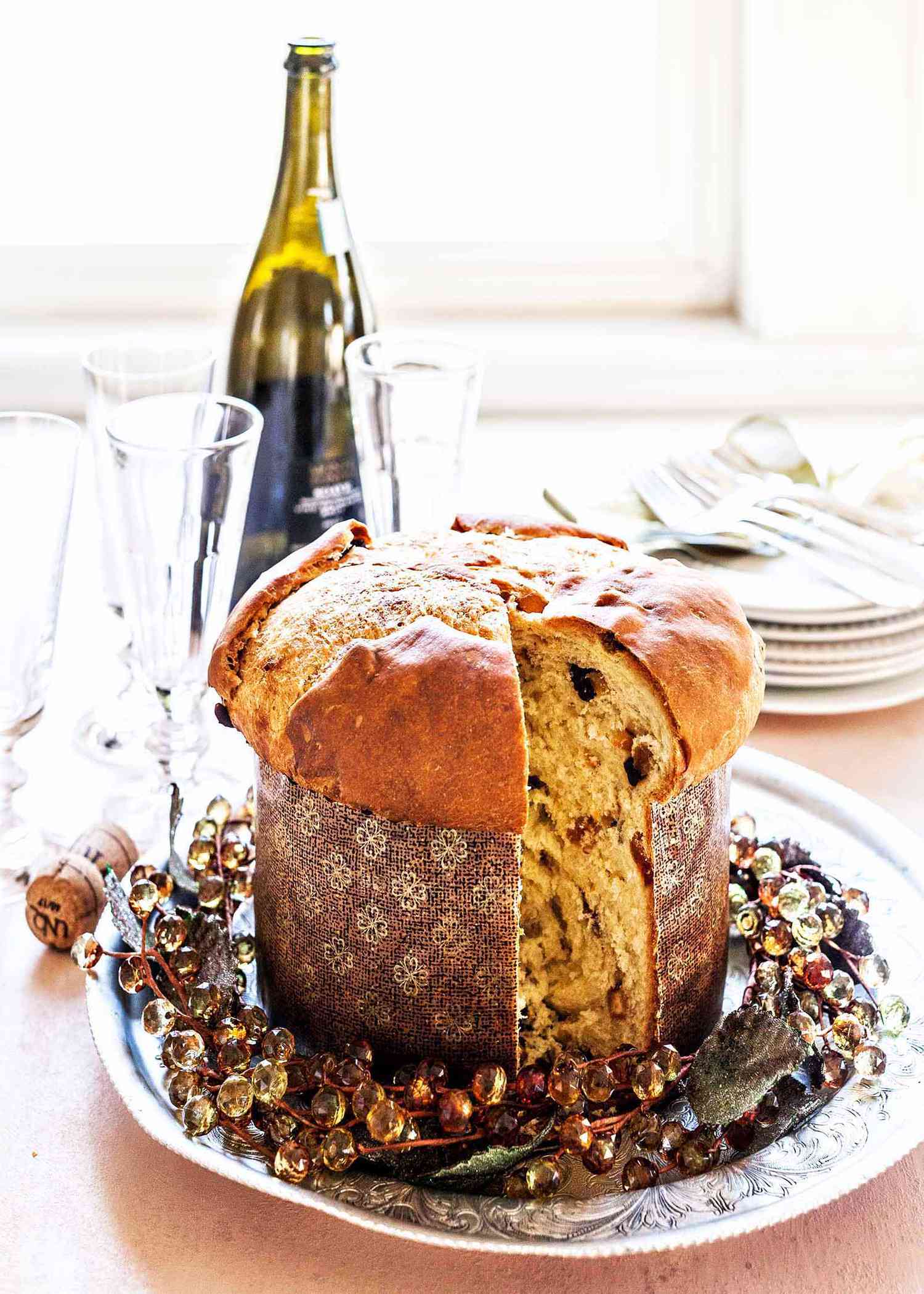 A loaf of homemade panettone in a paper mold with a slice cut out with a bottle of wine, and glasses in the background.