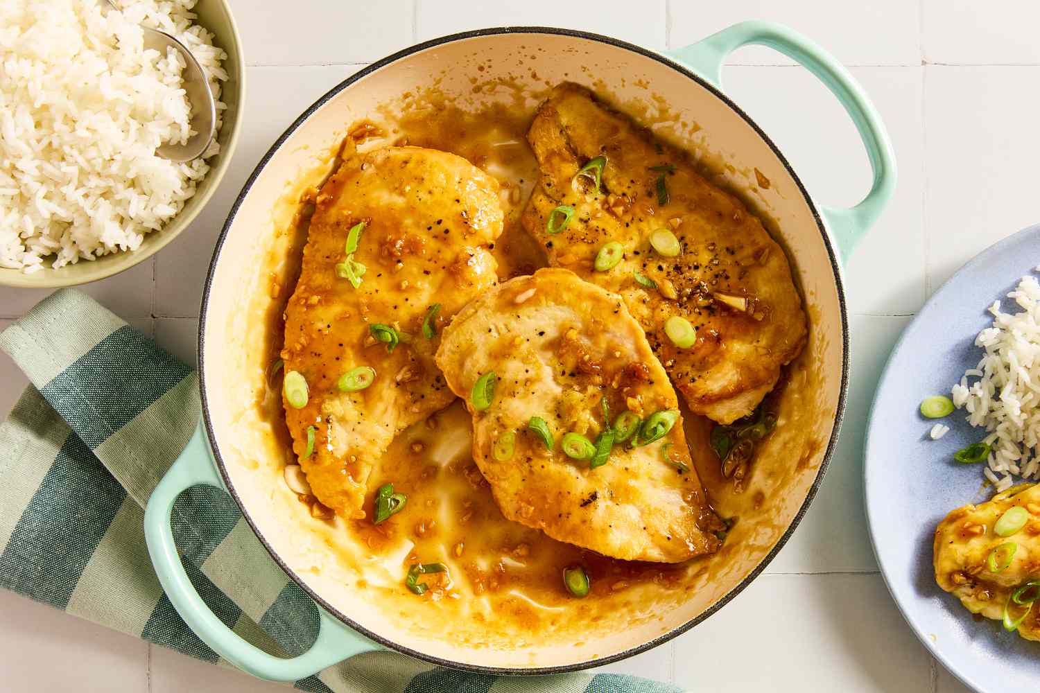 Overhead view of a teal dutch oven with three honey soy chicken breasts and sauce topped with green onions next to a bowl of white rice, a striped fabric napkin and blue plate of recipe and chicken