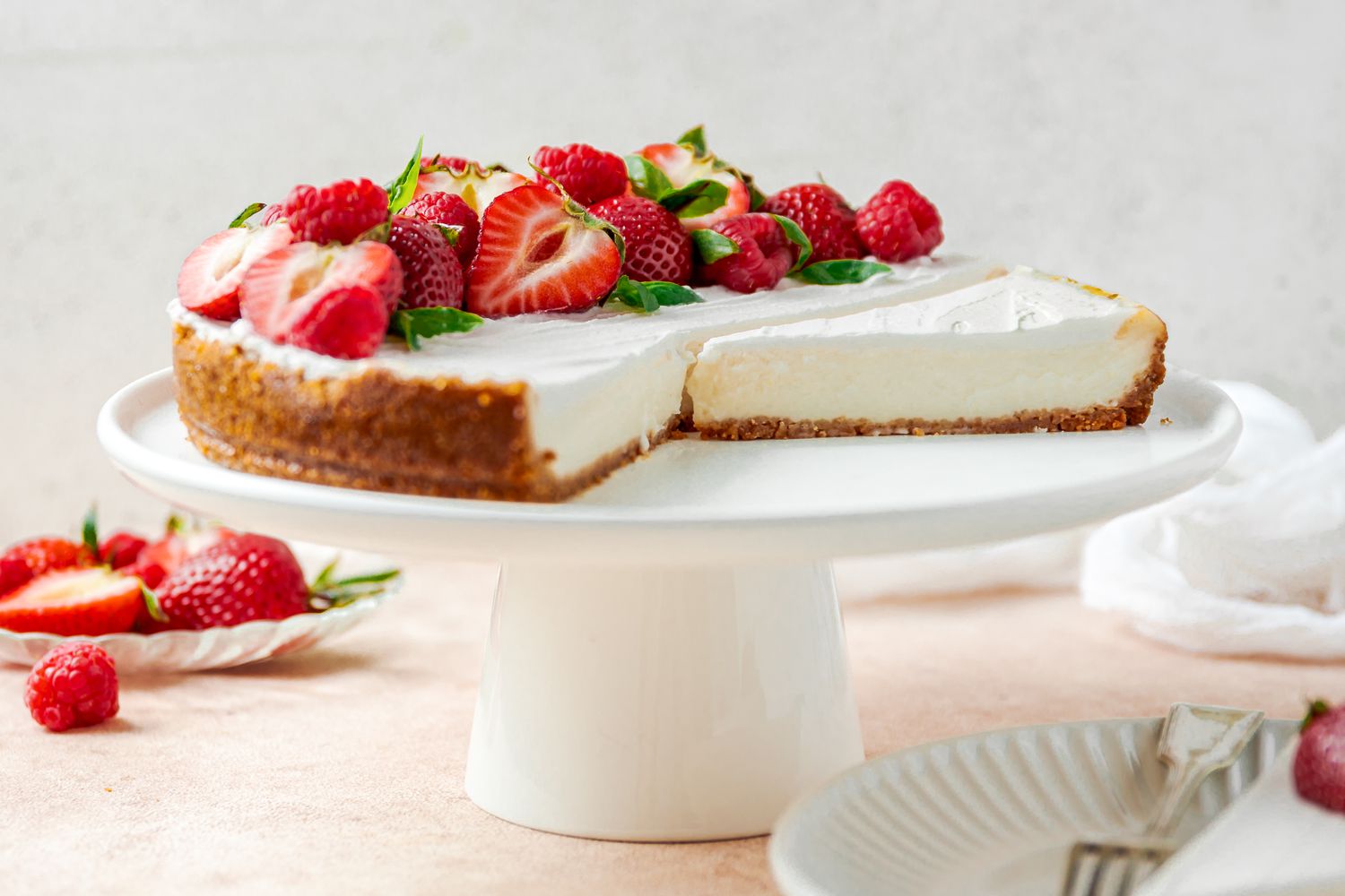 Vegan Cheesecake Topped With Halved Strawberries and Raspberries on a Cake Platter, and on the Counter Surrounding It, a Small Plate With More Fruit and Another Small Plate With a Slice of Cheesecake 