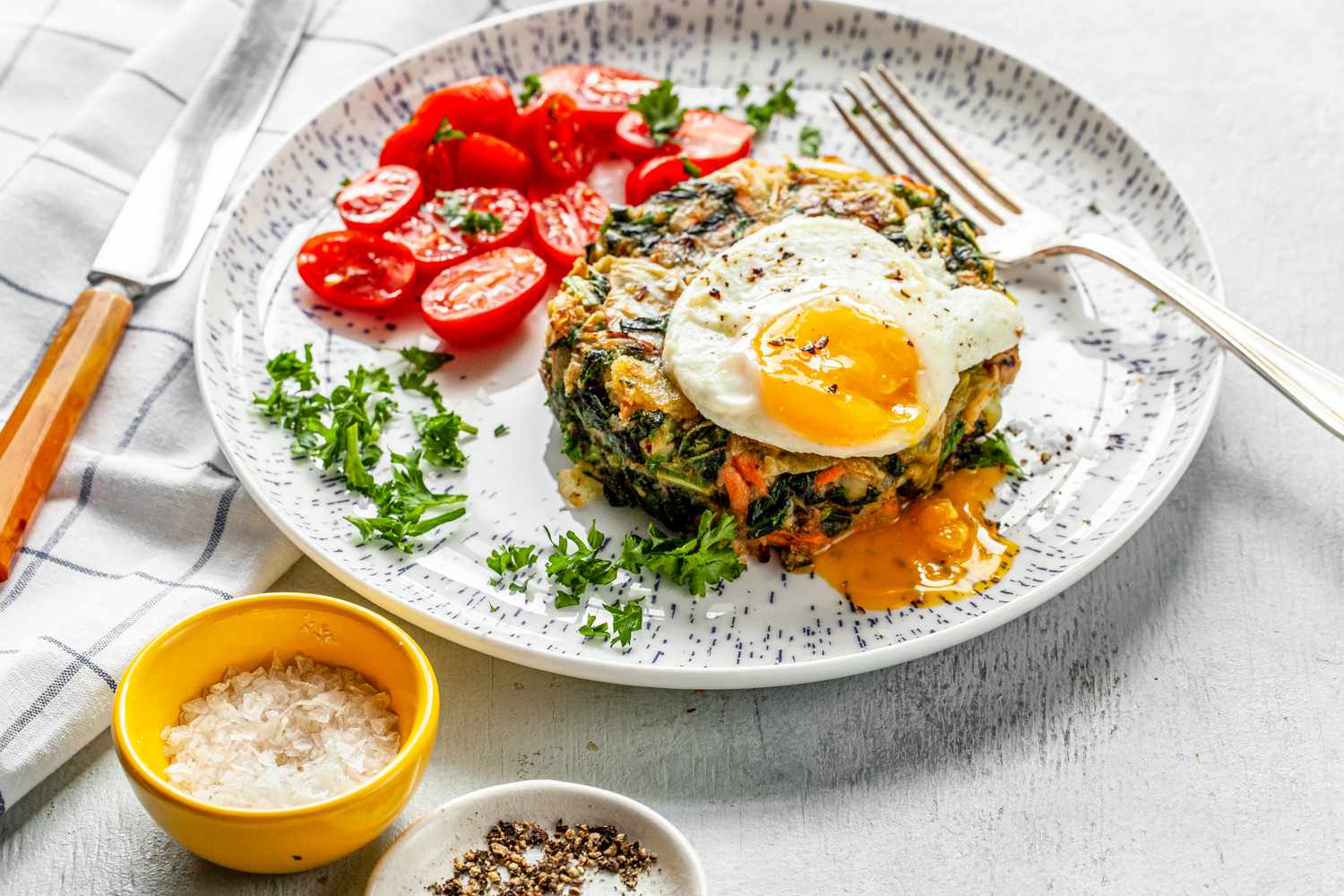 Bubble and Squeak Topped with Sunny Side Egg on a Plate with Halved Cherry Tomatoes and a Fork, Next to Bowls of Seasoning
