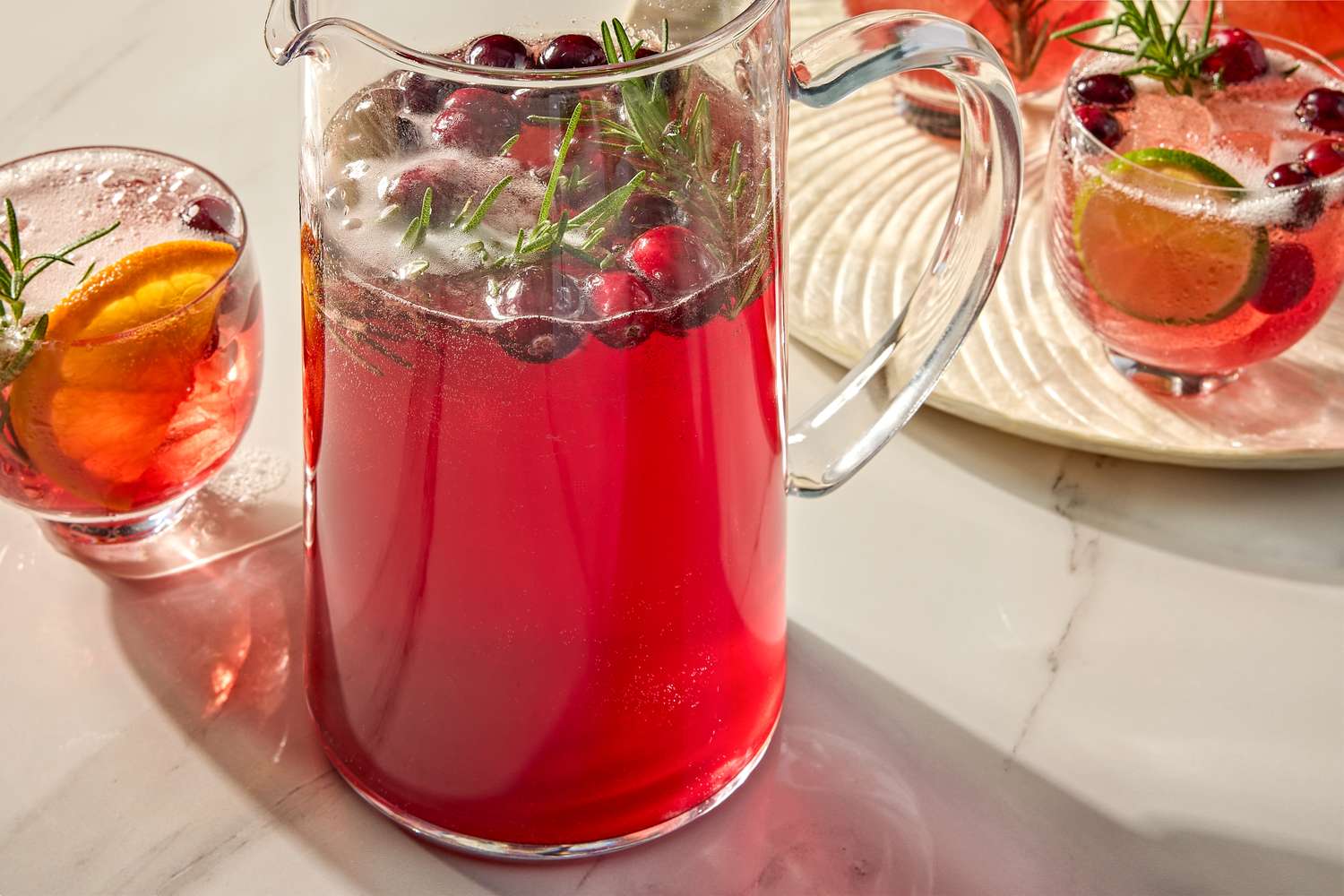 A pitcher of cranberry ginger spritz with garnished glasses nearby displayed on a table