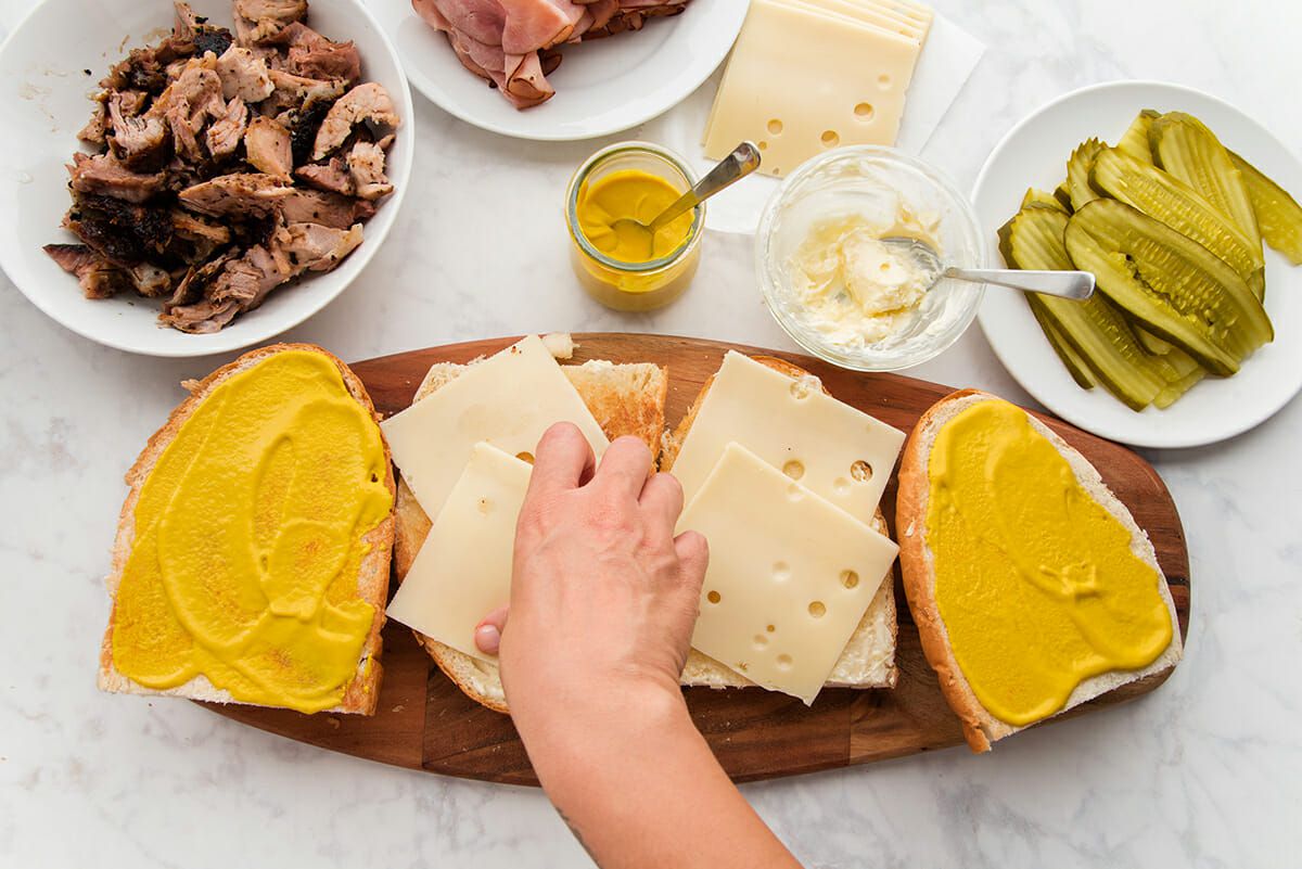A hand is placing sliced cheese on one quarter of a traditional cuban sandwich. One quarter has two slices of swiss cheese and two quarters are spread with yellow mustard. The sandwich is set on a wooden cutting board set on a marble table top. Bowls and plates of shredded pork, sliced ham, yellow mustard, sliced cheese, mayonnaise and sliced pickles are set above.
