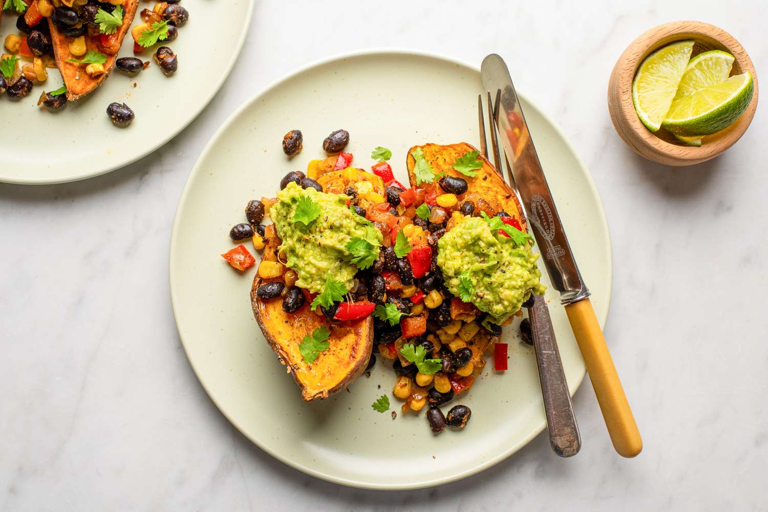 A dish featuring halved sweet potatoes topped with black beans vegetables guacamole and garnished with cilantro next to lime wedges