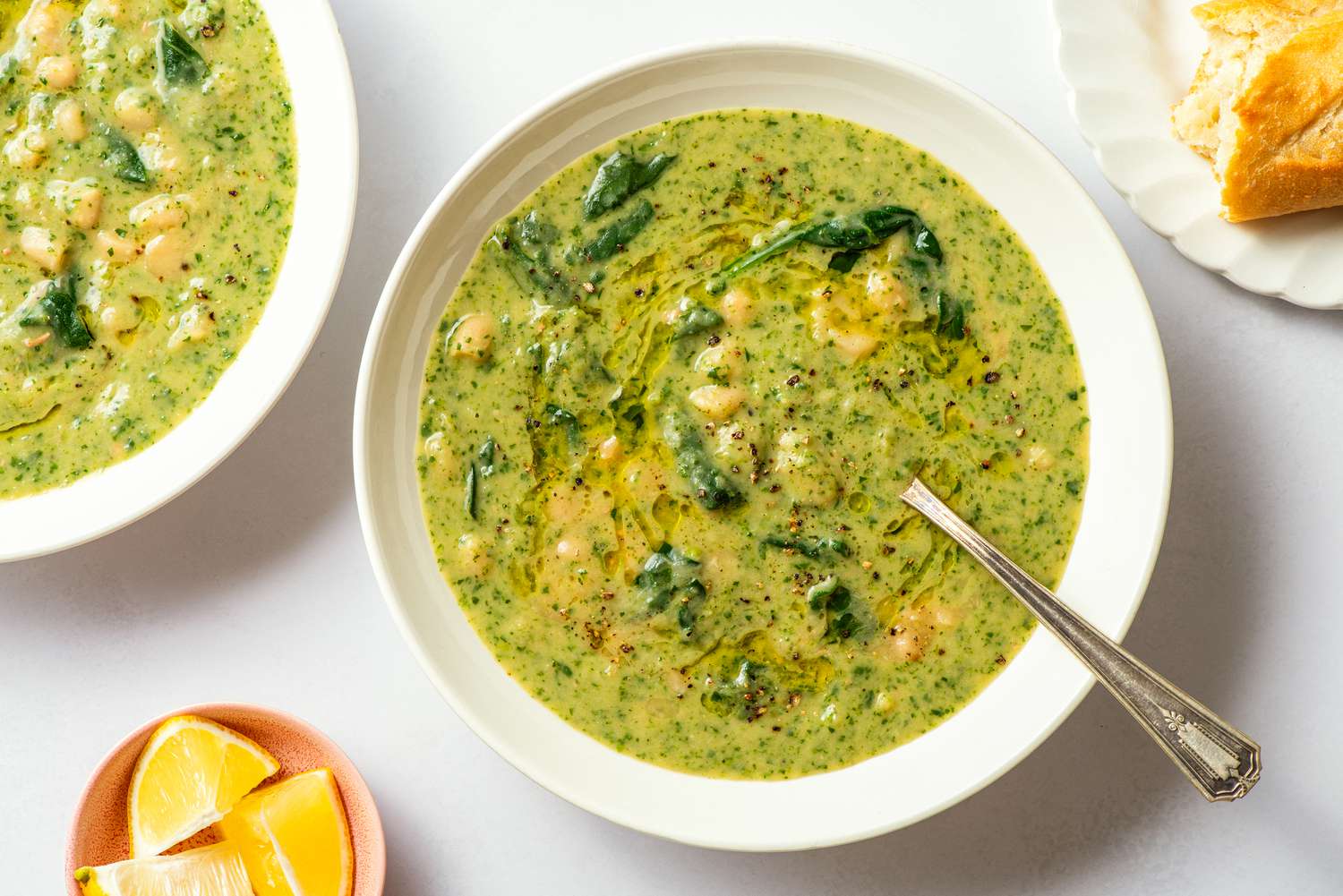 Overhead view of a white bowl of white bean and spinach soup with a spoon next to a plate of bread and bowl of lemon slices