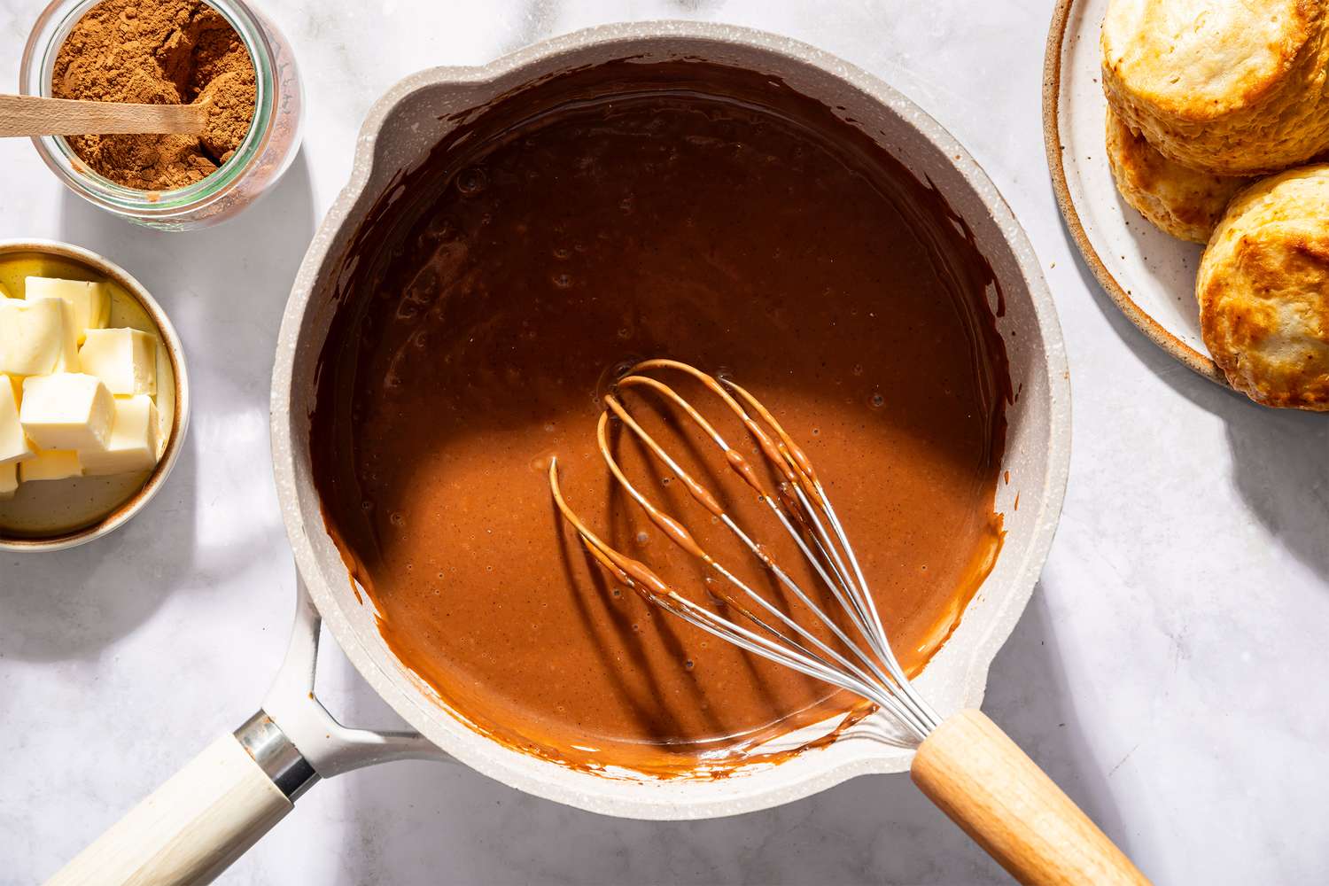 A whisk in a pot of chocolate gravy with butter and biscuits nearby