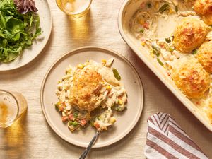 Serving of Chicken Cobbler With Cheddar Bay Biscuit Top on a plate at a table setting with a plate of salad greens, glasses of cider, and a casserole dish with cobbler