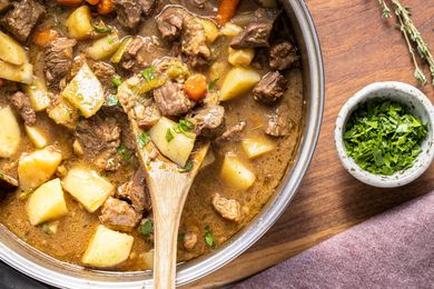 A pot of beef stew with carrots and potatoes a wooden spoon placed inside a bowl of herbs in the background