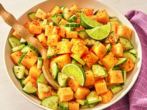 Bowl of cantaloupe pieces sliced cucumbers lime wedges and Tajn seasoning wooden spoon purple napkin nearby