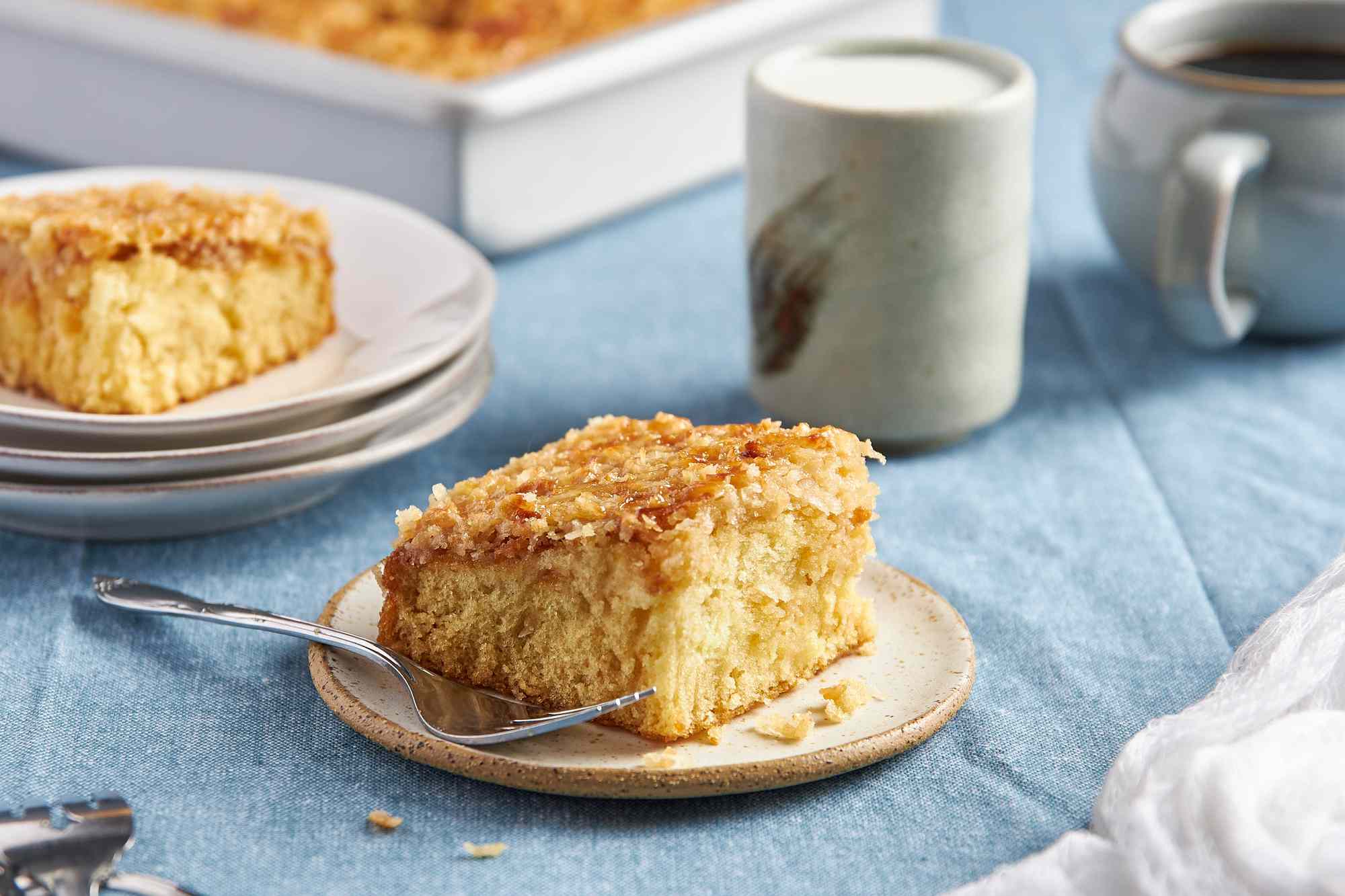 Slice of cake in a small plate with a fork, and in the surroundings, another slice on top of a stack of plates, a glass of cream, a mug of coffee, and a baking pan with more cake