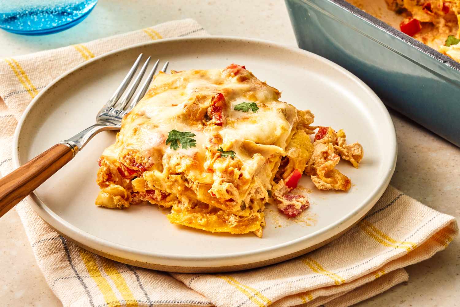 Side view of a beige plate with a slice of King Ranch Chicken Casserole and a fork, on top of a checkered napkin