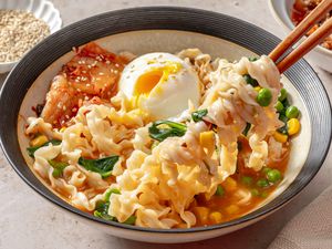 A bowl of noodle soup with an egg, kimchi, vegetables, and chopsticks lifting noodles, garnished with sesame seeds