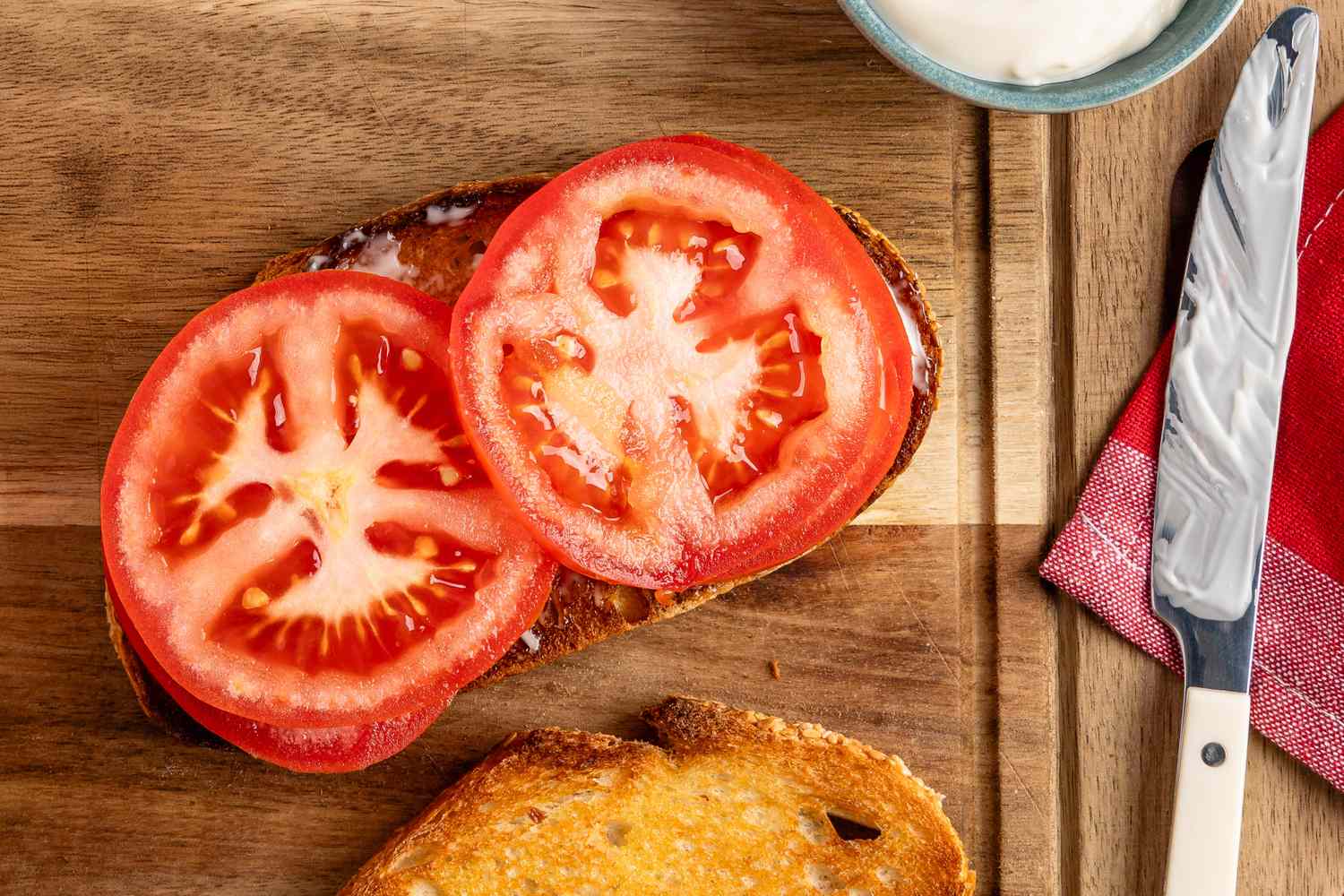 Open-faced tomato sandwich with sliced tomatoes on toast, accompanied by a knife and a small bowl of spread on a wooden surface