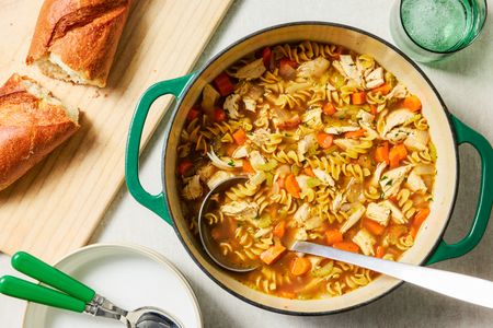 A pot of turkey soup with rotini pasta next to a cutting board with sliced baguette