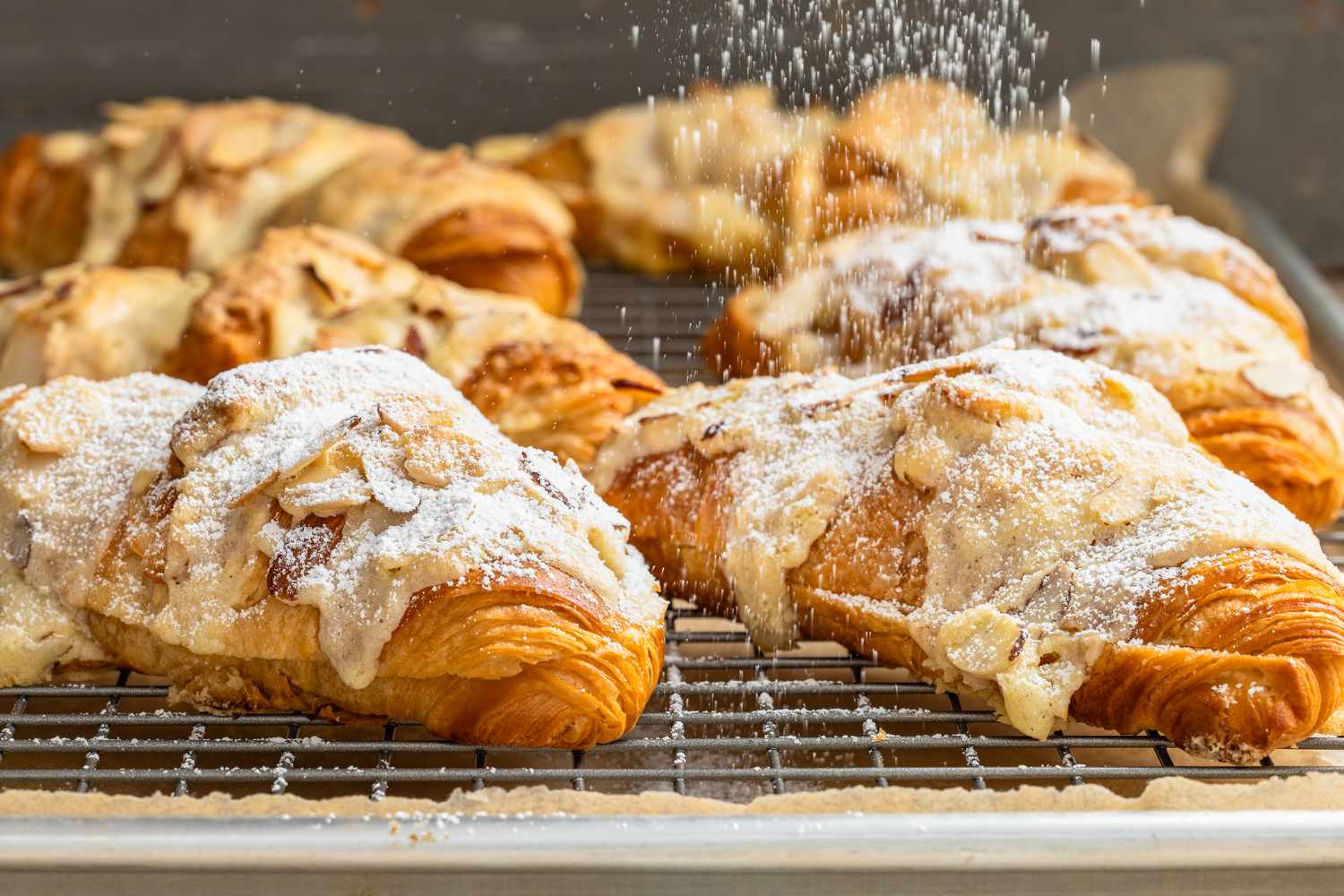 Almond Croissants Sprinkled With Powdered Sugar (Using a Colander) While on a Cooling Rack With a Piece of Parchment Paper Underneath It