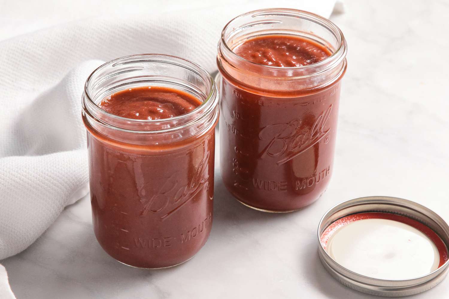 Two Mason Jars Filled With Banana Ketchup Next to a Lid and a White Kitchen Towel 