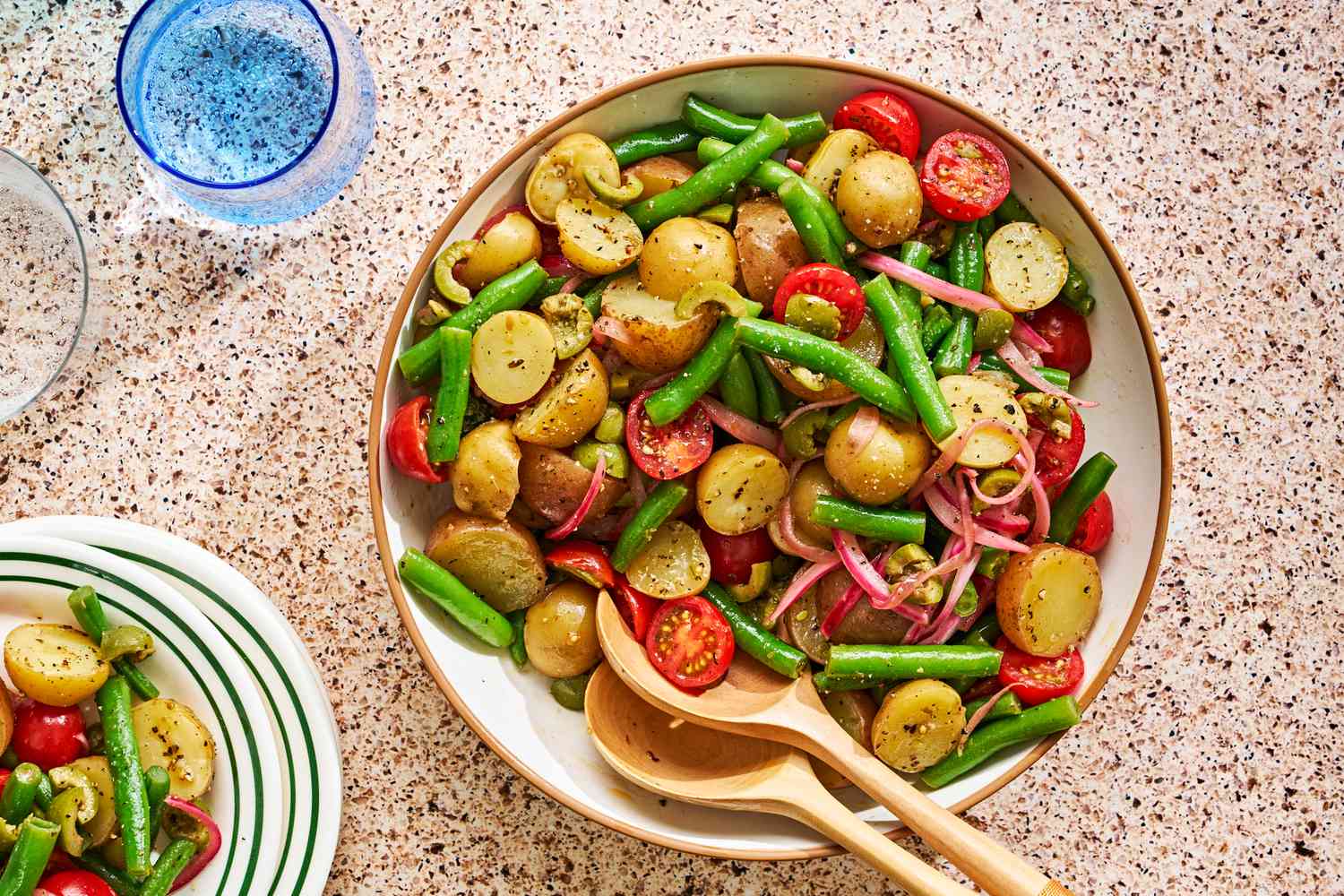 Sicilian potato salad in a bowl with serving utensils next to a glass of water and a stack of plates (with Sicilian potato salad on the top plate)