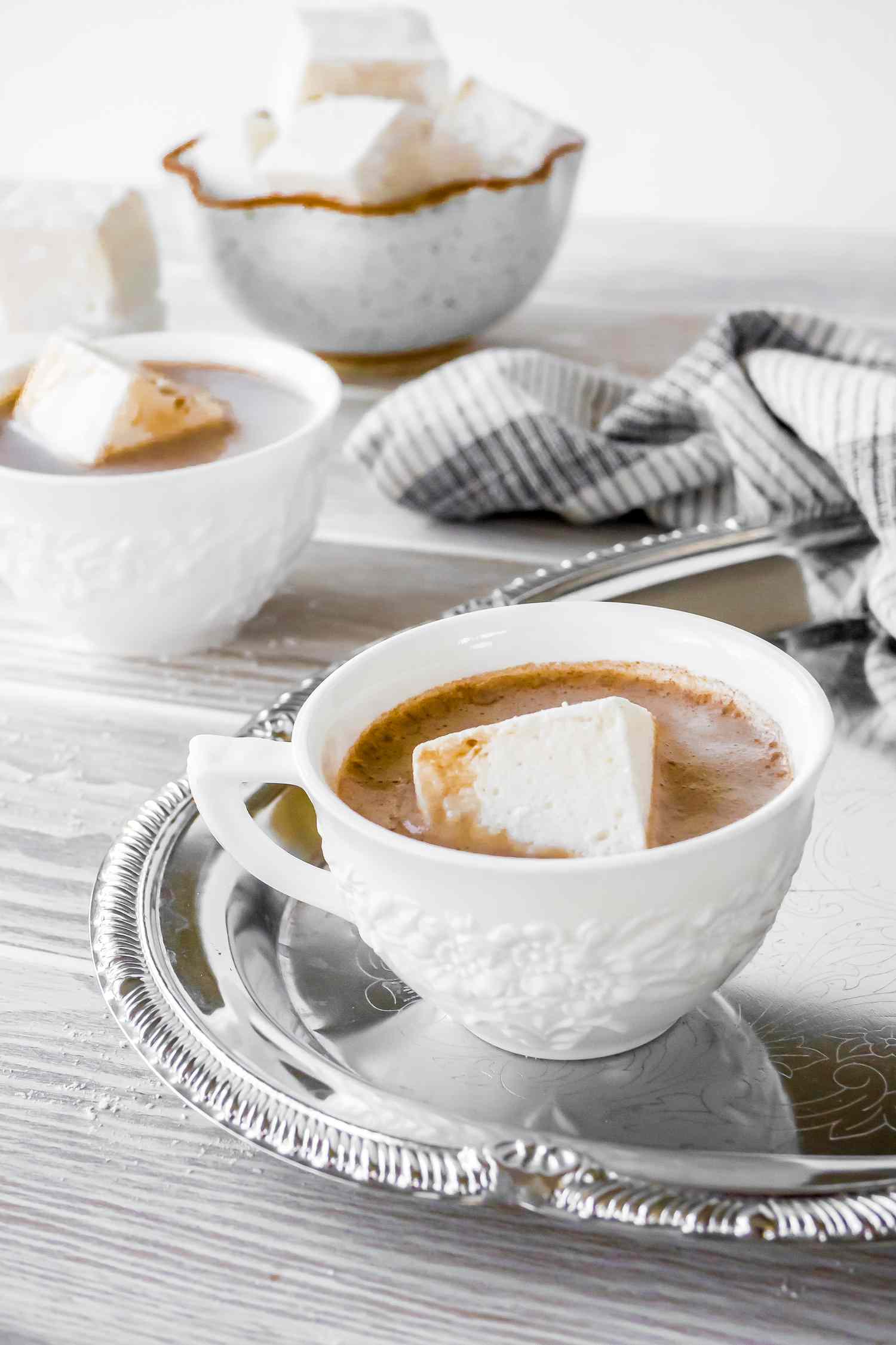 Cup of Hot Chocolate with a Large Vegan Marshmallow on a Tray with a Kitchen Towel. In the Background on the Counter Next to the Tray, Another Cup of Hot Cocoa with a Vegan Marshmallow and a Bowl with More Marshmallows.
