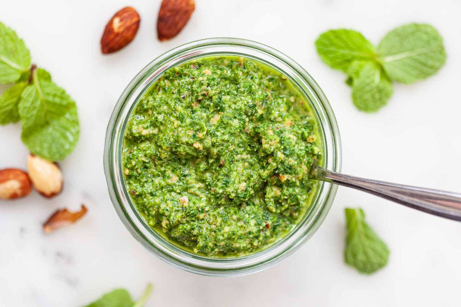 A jar of pesto with a spoon surrounded by some almonds and mint.