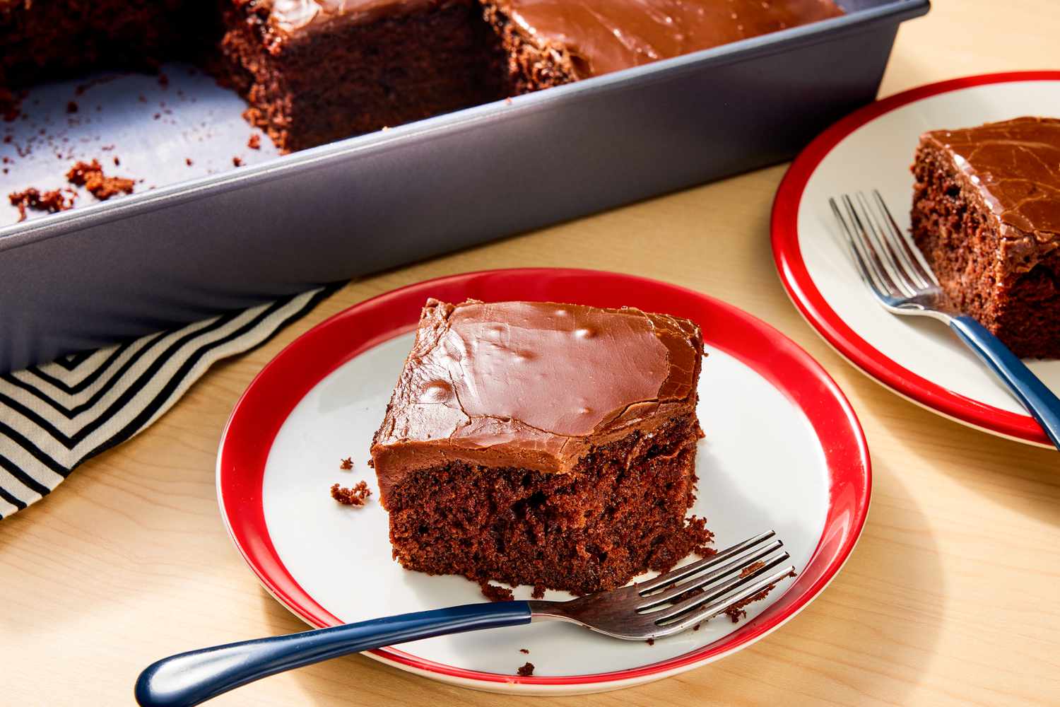 Two small plates of coca cola cake with forks in front of a metal baking pan of cake all on a wooden tabletop