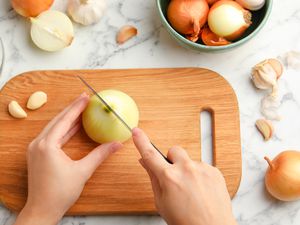 Overhead view of a woman's hands cutting an onion in half on a wooden cutting board, on a marble countertop next to other onions in a bowl and and garlic to the side