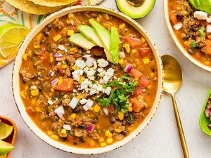Stovetop Taco Soup in a Bowl Topped with Avocado Slices, Cotija Cheese, and Cilantro and Surrounded by Bowls of Toppings