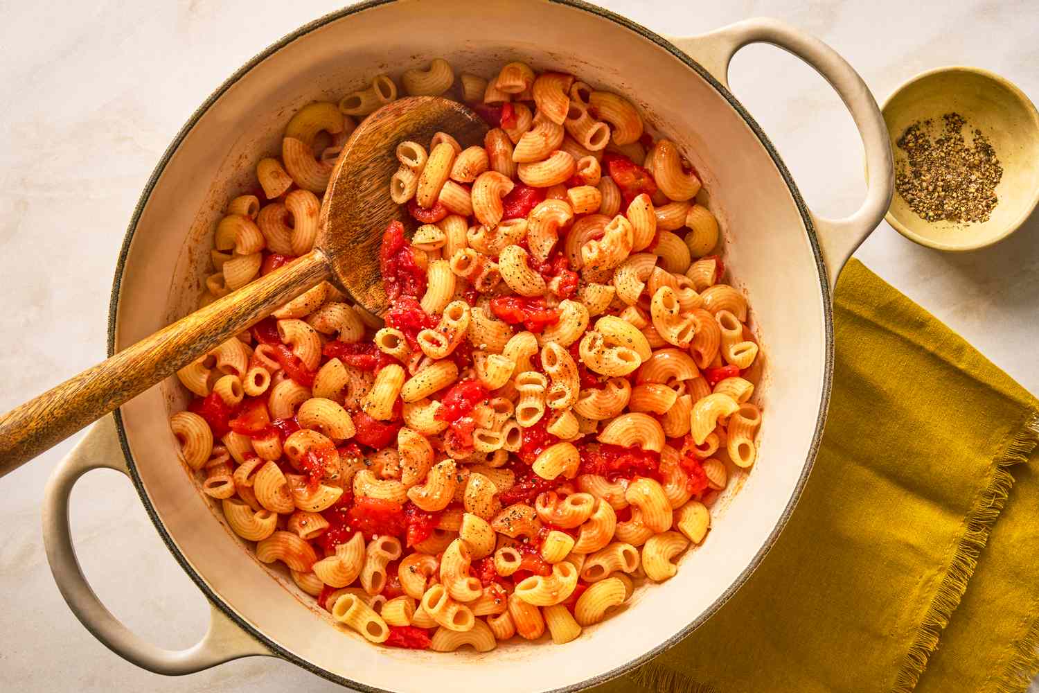 Pot of macaroni with a wooden spoon garnished with tomato pieces and pepper placed next to a small bowl and a yellow napkin