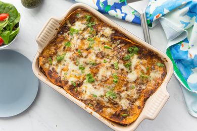 Beef Enchilada Casserole in a Casserole Dish Next to a Table Cloth, a Plate, and a Bowl with Side Salad 