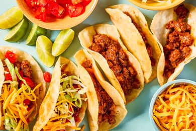 An assortment of tacos with beef shredded lettuce shredded cheese garnishes and lime wedges