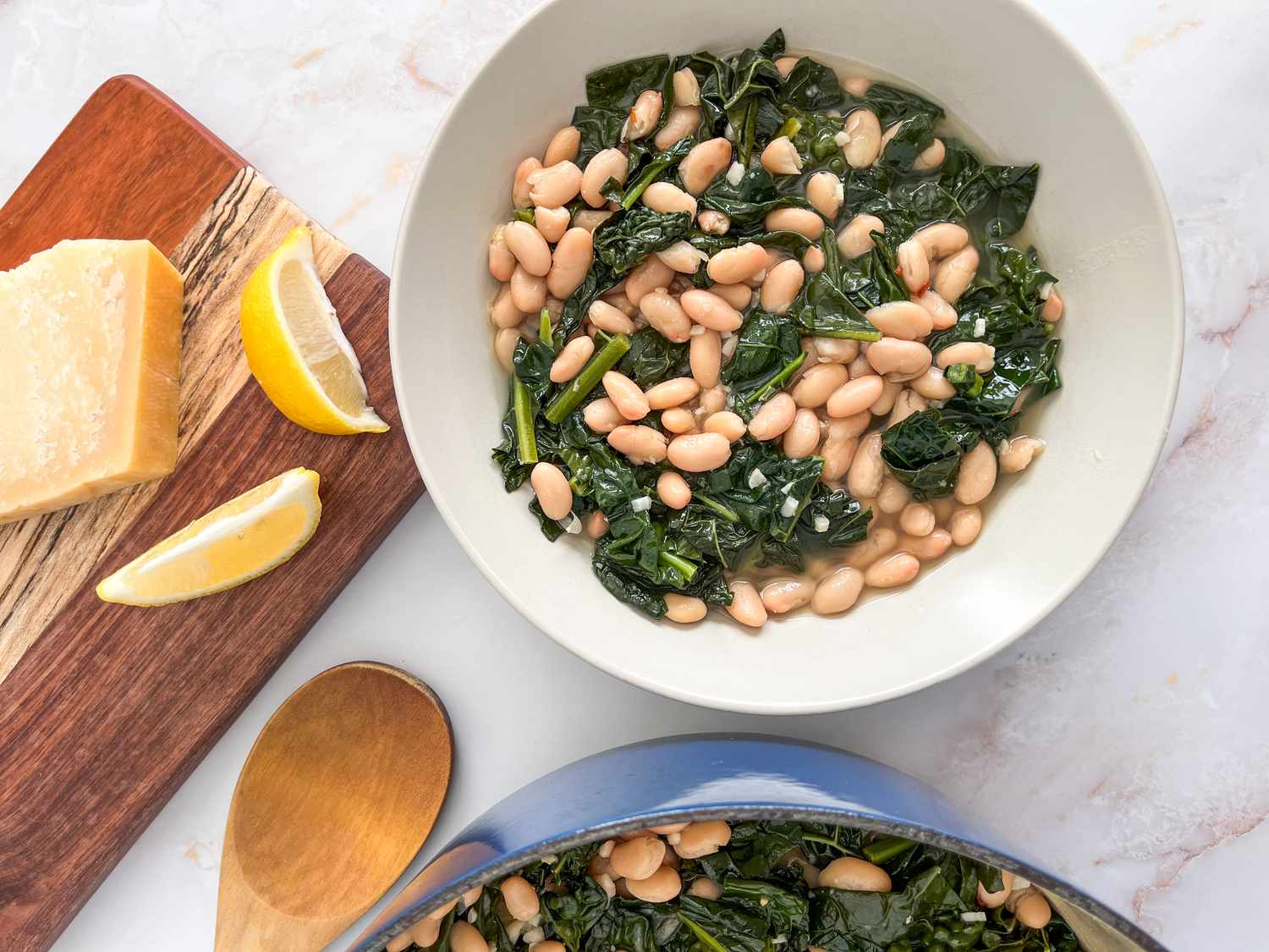 Bowl of Italian beans and greens next to a pot with more, a wooden board with lemon wedges, and a block of parmesan