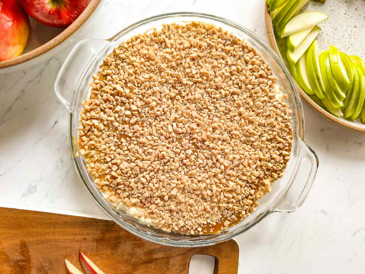 Glass dish filled with a caramel apple dip topped with chopped nuts surrounded by sliced apples on a wooden board and a plate