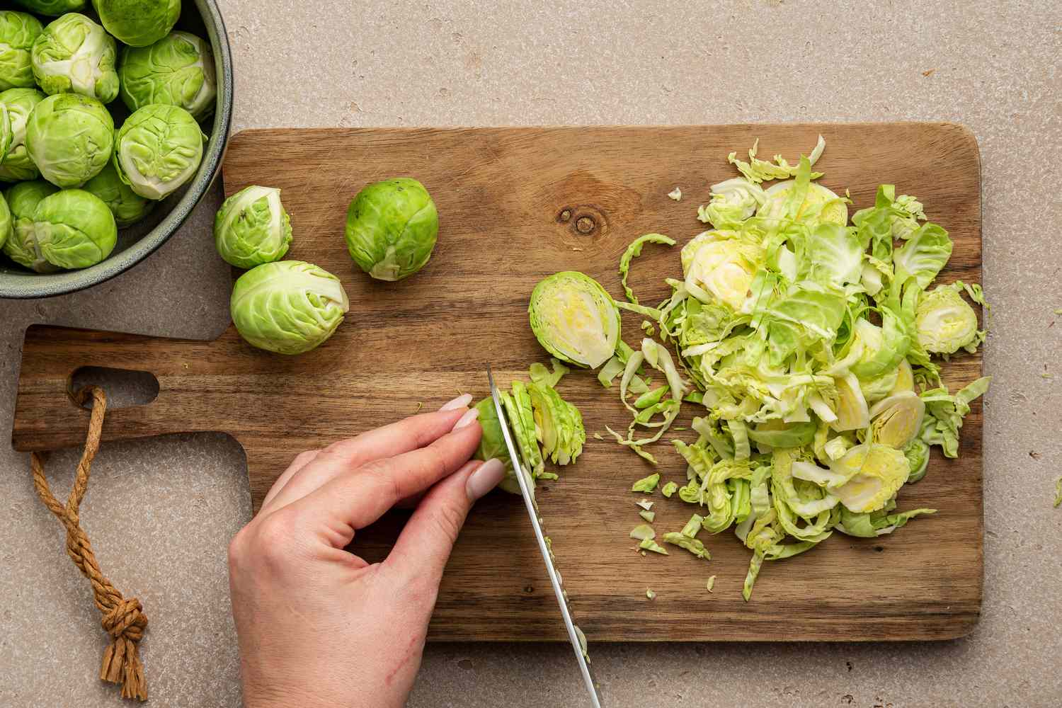Brussels Sprouts Cut into Thin Slices on a Cutting Board Using a Knife Next to a Bowl of Whole Brussels Sprouts 
