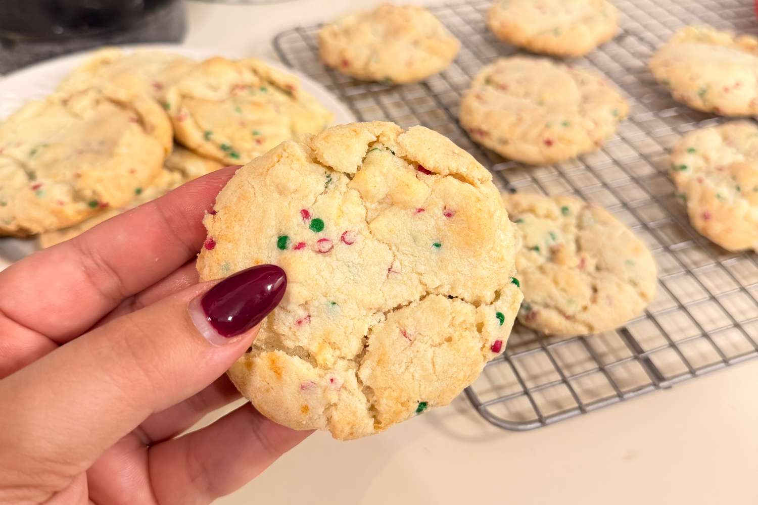 A hand holds a cookie with colorful sprinkles with more cookies on a cooling rack in the background