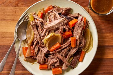 Overhead view of a plate of pot roast with carrots and onions and a fork and spoon all next to a cup of gravy on a wooden tabletop