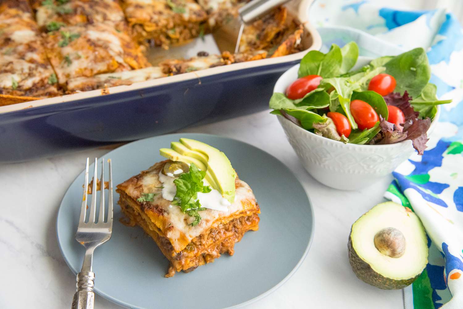 Plate with a Slice of Beef Enchilada Casserole Topped with Sour Cream, Cilantro, Jalapeno, and Sliced Avocado, Surrounded by a Half Cut Avocado, a Bowl with Side Salad, a Table Cloth, and a Casserole Dish with More