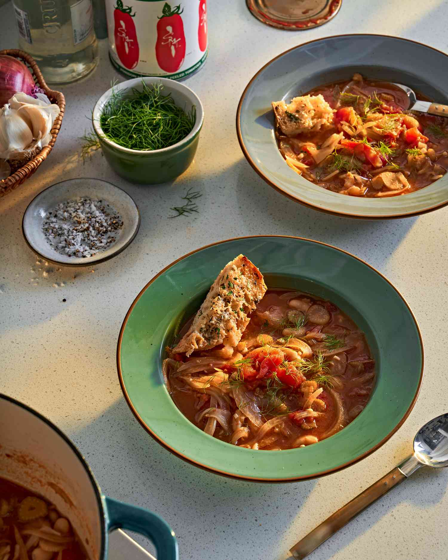 Two bowls of tomato, white bean, and fennel stew on a kitchen counter with side bowls of salt and pepper and fennel fronds